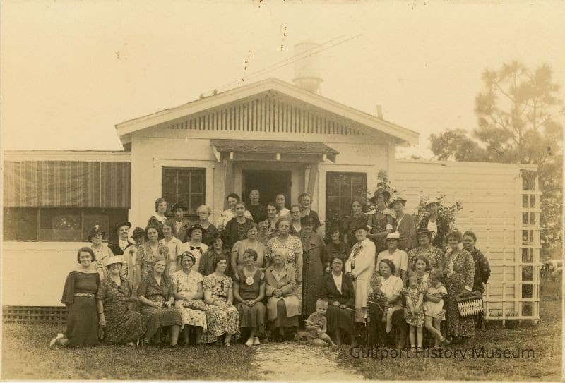 A black and white photograph postcard of the Home Demonstration Club posing in front of 5314 Lakeview Ave (5314 Gulfport Blvd). All of the members of the club are women in various day dresses and headwear. Four small children are at the front of the group. A water tower is visible in the far background above the house.