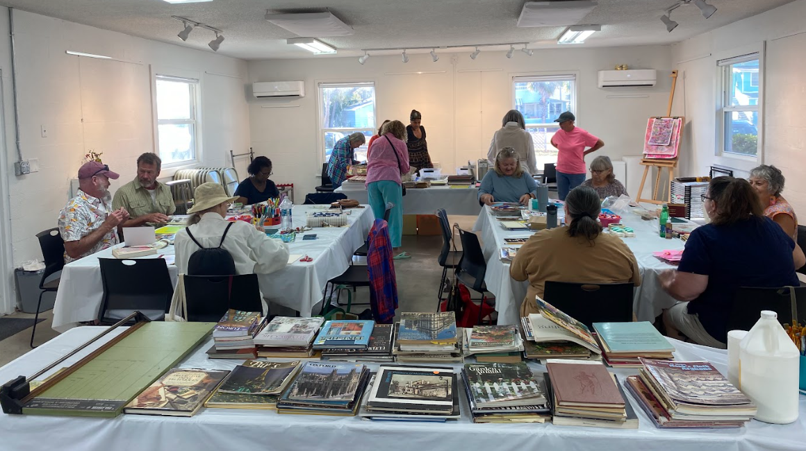 A picture of a large group of people working on art around 3 tables, with a 4th table covered in book in the forefront