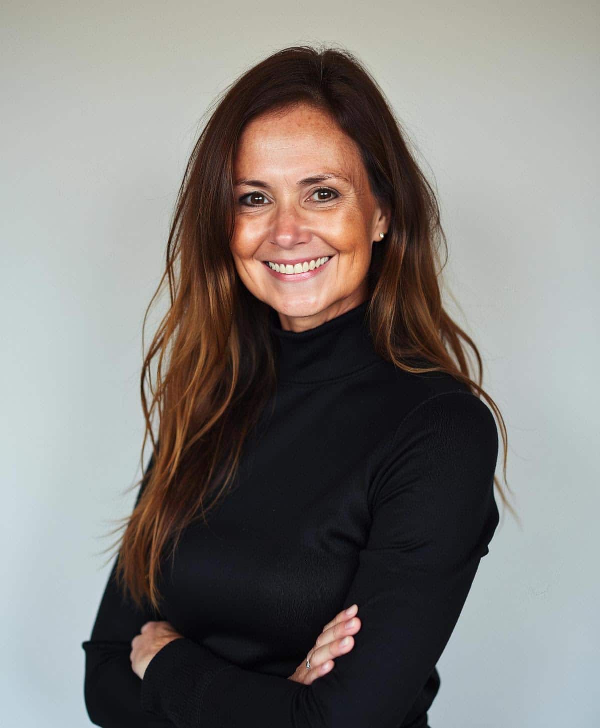 Smiling woman. Long straight brown hair. Black turtleneck. Arms crossed. Plain light background.