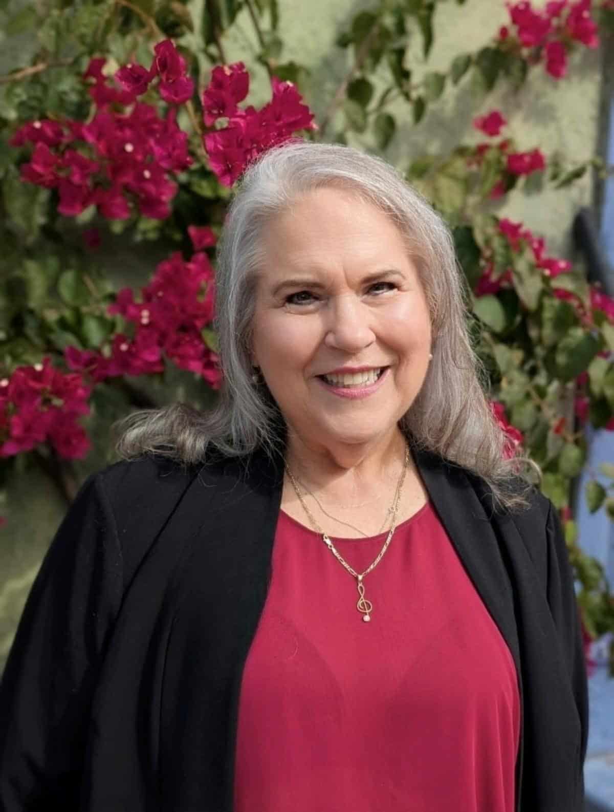 A smiling woman with long gray hair. Wearing a black jacket and a red blouse stands in front of vibrant pink flowers.