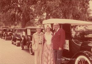 A color photograph of Frederick Allen, Yvonne Johnson, and Bert Williams standing in front of a row of antique cars at the Citizens Parade. Yvonne is wearing period costume.