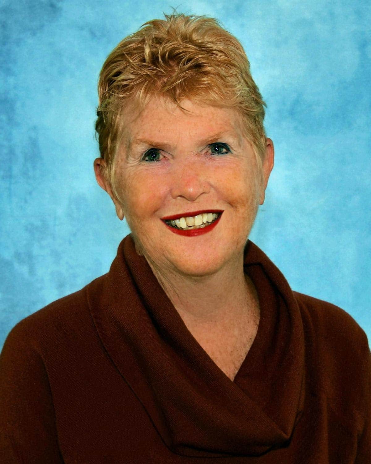 Smiling woman with short reddish hair wearing a brown top against a light blue background.